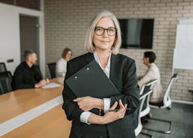 Senior businesswoman with eyeglasses holding a clipboard in a modern conference room with colleagues.