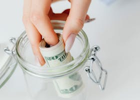 A close-up of a hand placing rolled dollars into a glass jar, symbolizing savings.